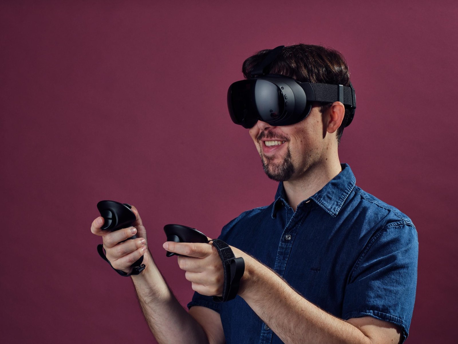 Sean Hollister smiles as he wears the Steam Frame, a small black cordless headset with a highly reflective front face. He holds two wands that are strapped around his wrists in front of him, with visible triggers and bumpers and saucer-shaped tops. 