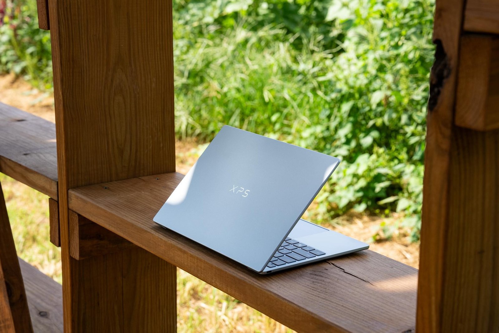 A sky colored Dell XPS 13 (2026) laptop sitting on a wood surface with bright sunlit greenery in the background.