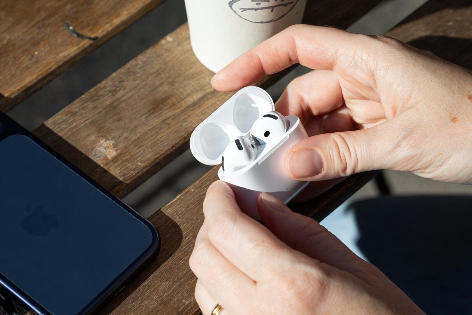 Hands holding the Apple AirPods 5 earbuds above a wooden-slated table with an iPhone and coffee cup on it.