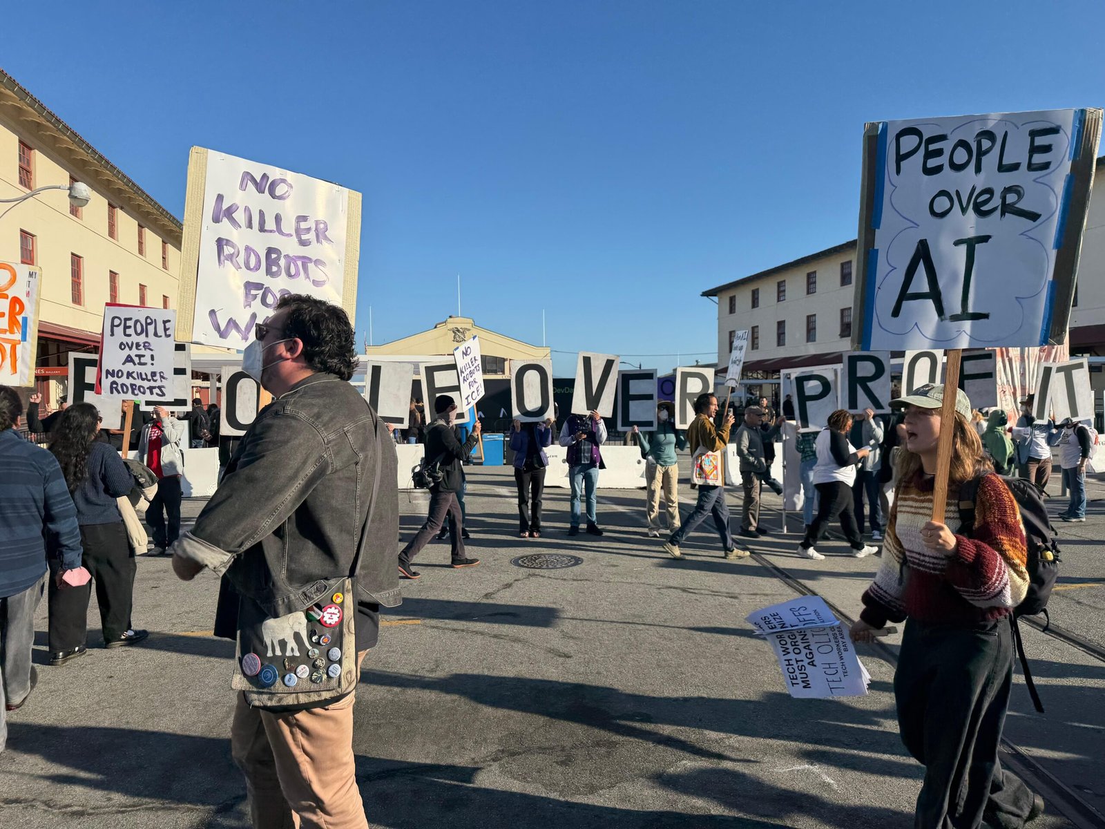 Protesters holding signs with messages like “People over AI” congregating outside an event venue.