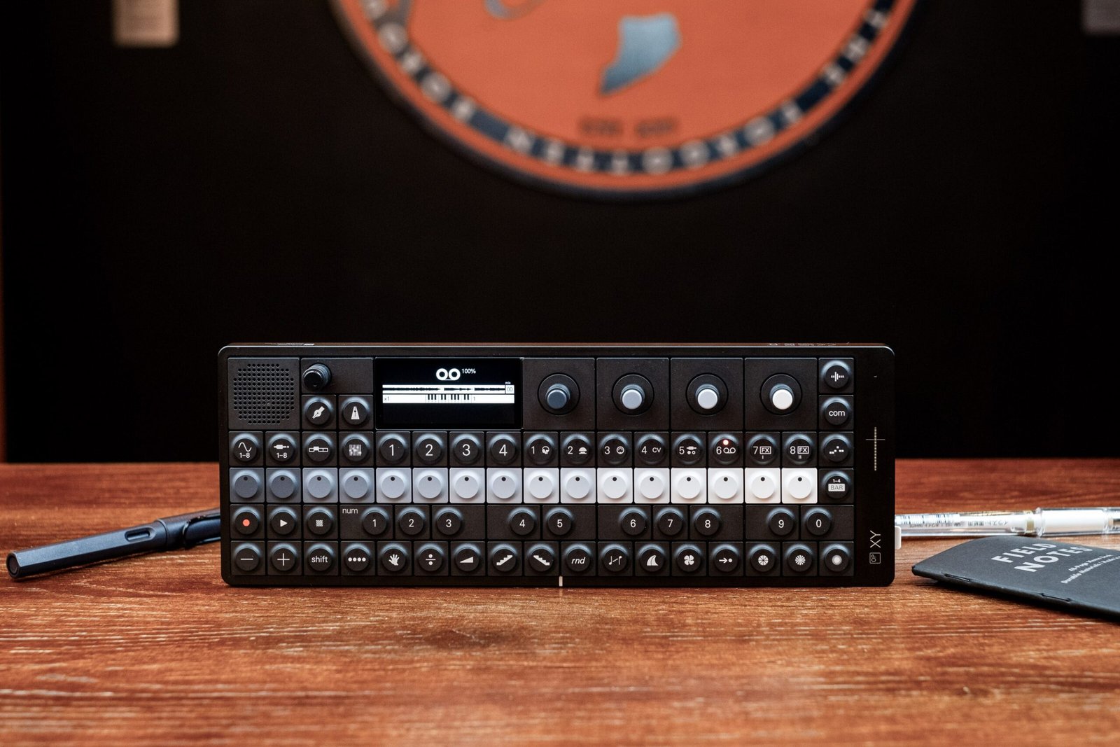 A black-and-grey rectangular device standing on its front edge on a wooden table. It has rows of buttons along the bottom half of the front face; the top half has a small LCD screen and several knobs. It’s an electronic musical instrument about half the size of a computer keyboard.