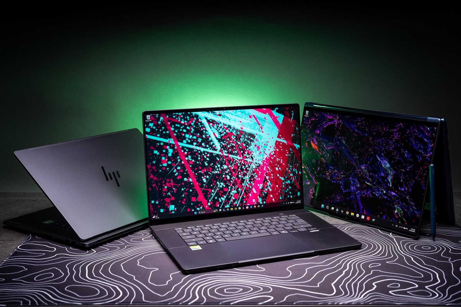 RTX Spark versions of the HP OmniBook Ultra (left), an Asus ProArt P16 (center), and a Lenovo Yoga 9n (right) on a dark gray table against a green-illuminated backdrop.